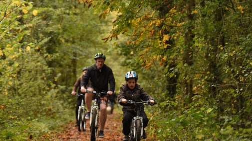 A family wearing helmets and coats cycles along the Tramway Trail at Calke Abbey, beneath a canopy of autumnal leaves
